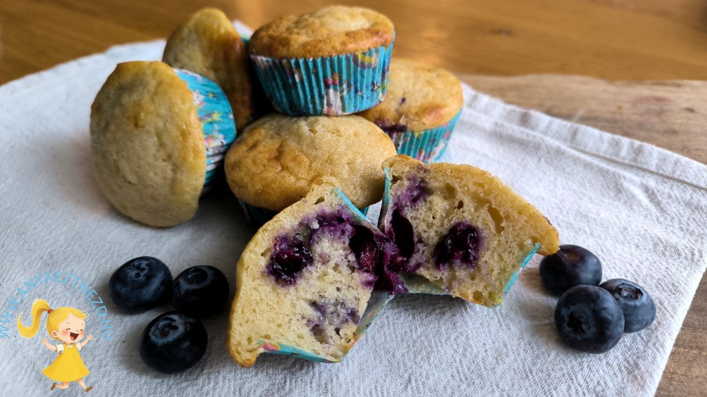 Kék áfonyás muffinok egy fehér szalvétán, néhány kék áfonyával az előtérben, az egyik muffin felvágva, belül áfonyadarabokkal.