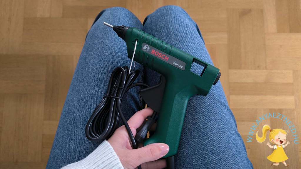 Egy Bosch kézi ragasztópisztoly, zöld színben, látható egy személy kezében, aki farmernadrágot visel.