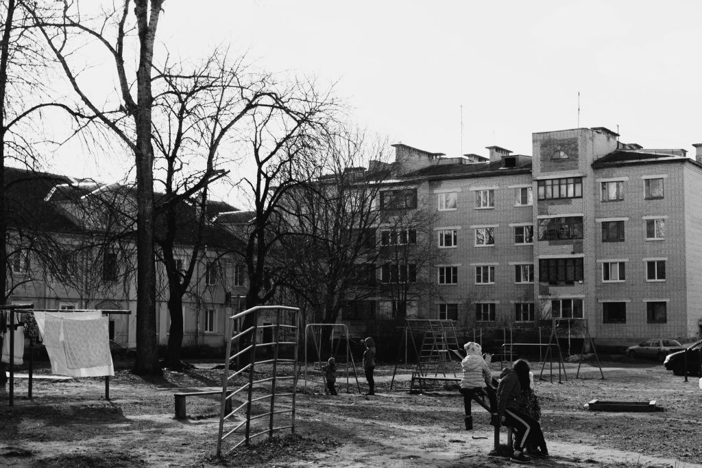 Fekete-fehér kép egy játszótérről, ahol gyerekek játszanak hinták és mászókák mellett, háttérben a fák és lakóépületek láthatók.