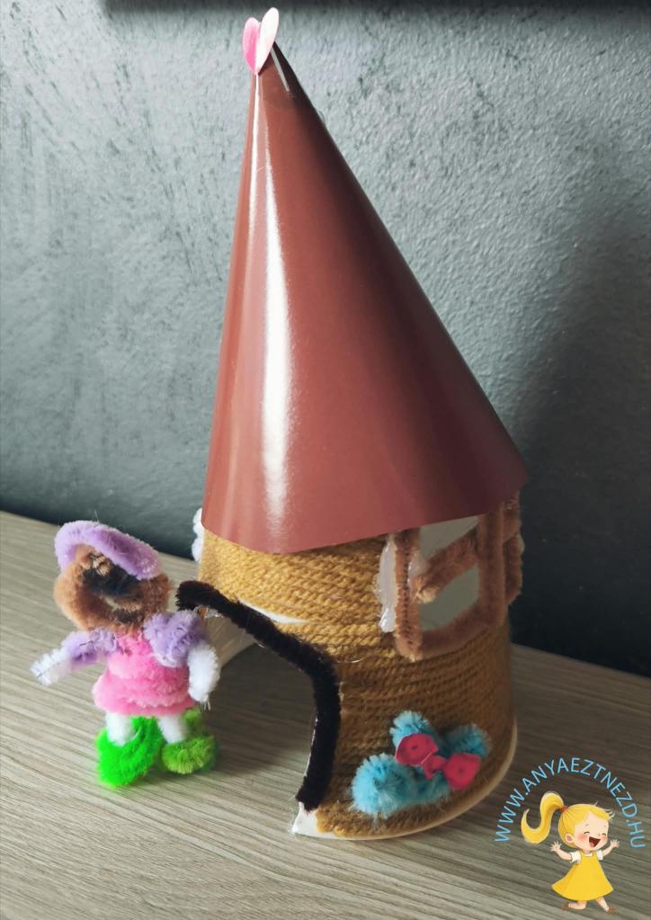 Egy kézműves házikó, amely fonallal és színes pipatisztítóval készült, mellette egy kis figura áll, aki vidám színű ruhát visel.