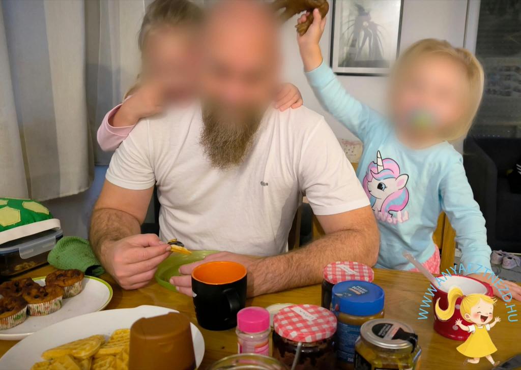 Egy férfi ül az asztalnál, miközben két gyerek játszik körülötte. Az asztalon különféle ételek, édességek és italok találhatók.