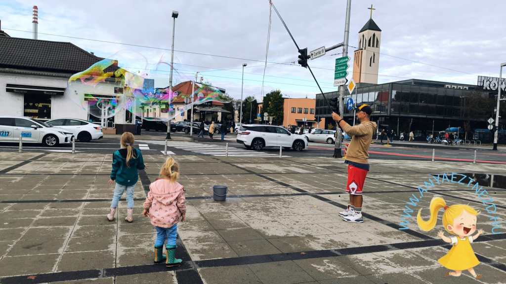 Két gyermek játszik a tér közepén, miközben egy felnőtt hatalmas buborékokat fúj. A háttérben épületek és autók láthatók, a város utcáján.