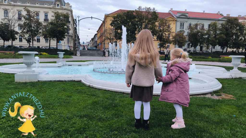 Két kislány áll egy szökőkút előtt, zöld fűben, a háttérben városi épületek láthatók.