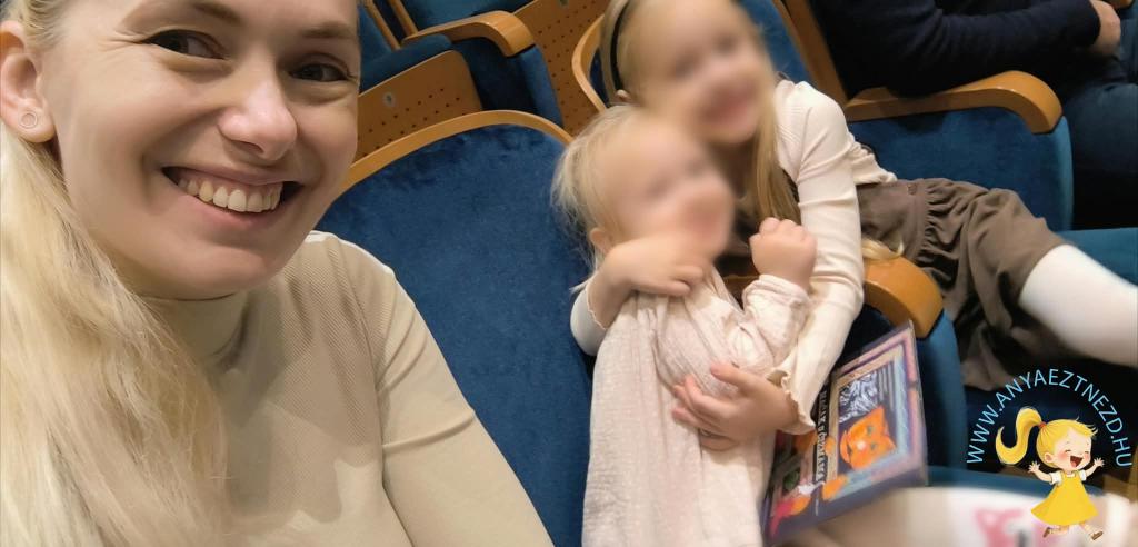 Egy nő mosolyog a kamerába, miközben két kisgyerek ölelkezik mellette, színházi környezetben.